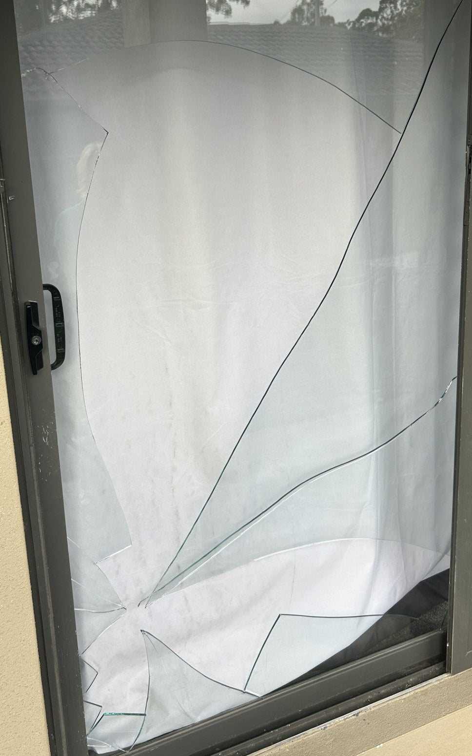 A damaged sliding glass door screen with a large section of shattered glass and a torn white curtain, set against a beige exterior wall.